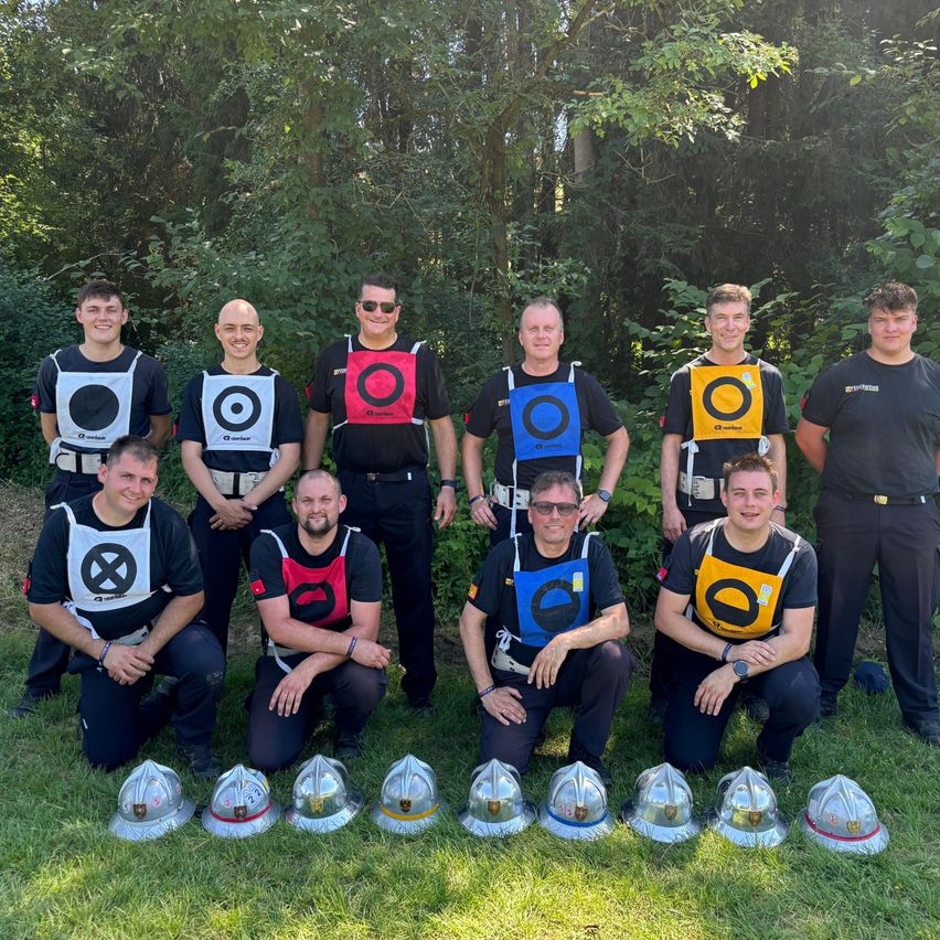 Eine Gruppe Feuerwehrleute posiert für ein Foto im Wald, jeder trägt eine nummerierte Weste und steht auf einem Rasen mit davor aufgestellten Helmen.