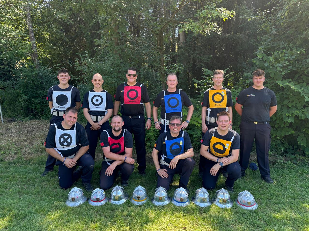 Eine Gruppe Feuerwehrleute posiert für ein Foto im Wald, jeder trägt eine nummerierte Weste und steht auf einem Rasen mit davor aufgestellten Helmen.