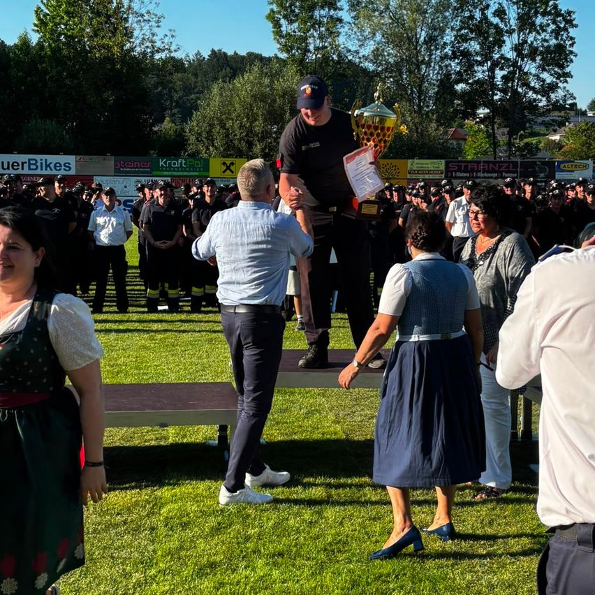 Ein Mann steht auf der Bühne und hält einen Pokal, während andere zuschauen, einschließlich Leute in Uniformen, an einem sonnigen Tag.