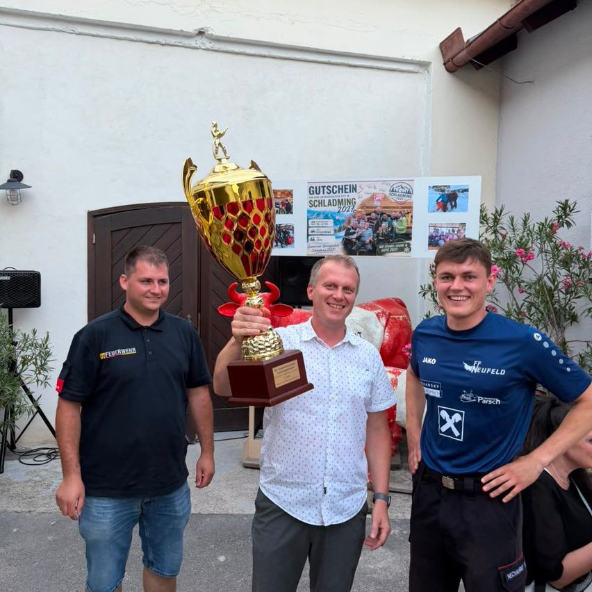 Drei Männer stehen vor einem Gebäude, einer hält eine goldene Trophäe und lächelt. Der Mann auf der linken Seite trägt ein schwarzes T-Shirt mit Text und Jeans. Der Mann in der Mitte trägt ein weißes Poloshirt und graue Hose. Der Mann auf der rechten Seite trägt ein blaues T-Shirt und schwarze Hose. Hinter ihnen ist ein Schild angebracht.
