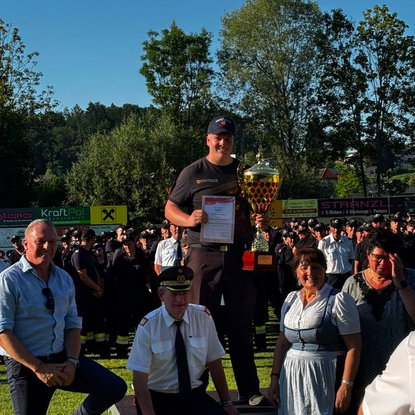 Ein Mann in Uniform hält einen Pokal und ein Zertifikat in der Hand, während er auf einer Plattform steht. Dahinter stehen weitere uniformierte Personen in einer Reihe. Zwei Frauen und ein Mann stehen in der Nähe. Bäume und Banner sind im Hintergrund.