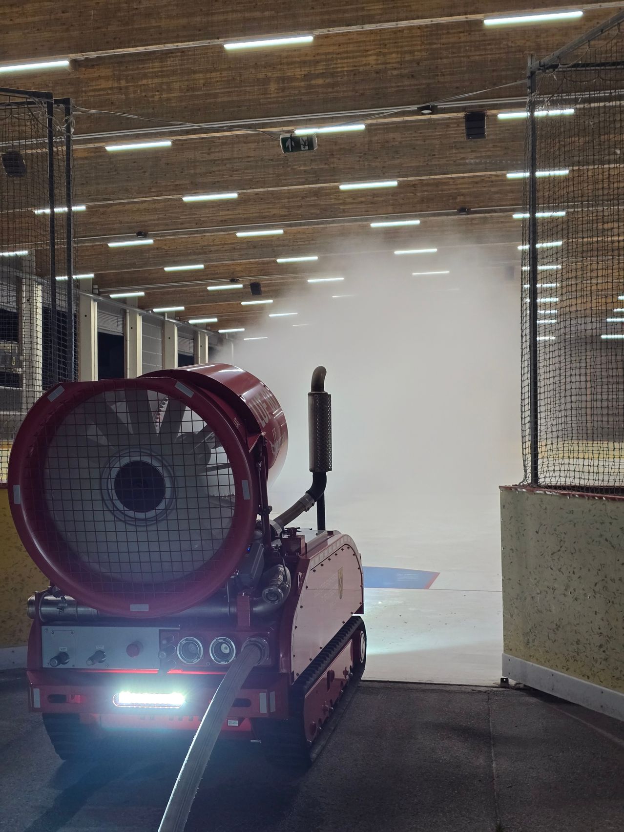 Eine überdachte Eisbahn mit einer roten Maschine, die Dampf abgibt. Die Maschine steht auf dem Boden in der Nähe einer Wand. Die Eisbahn ist von einem Netz umgeben.