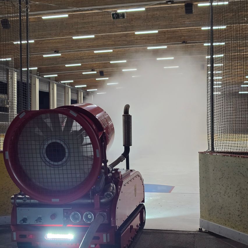 Eine überdachte Eisbahn mit einer roten Maschine, die Dampf abgibt. Die Maschine steht auf dem Boden in der Nähe einer Wand. Die Eisbahn ist von einem Netz umgeben.
