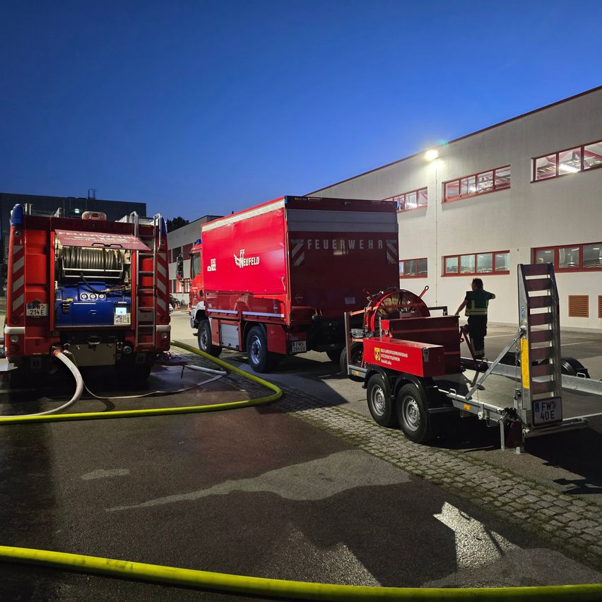 Zwei Feuerwehrfahrzeuge sind in der Nähe eines Gebäudes geparkt. Ein Fahrzeug hat eine Leiter und einen Schlauch an der Seite. Das andere hat einen Anhänger angeschlossen.