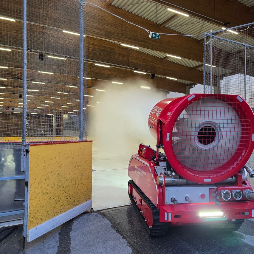 Eine rote Maschine mit einem großen Lüfter stößt Wasserdampf in einer Sporthalle mit Metallzäunen und einer gelben Barriere aus.