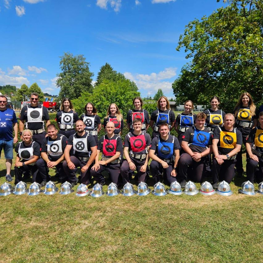 Eine Gruppe von Menschen in Sicherheitswesten mit Zieldesigns posiert für ein Foto auf einem Grasfeld. Vor ihnen stehen mehrere silberne Helme. Dahinter sind Bäume und ein blauer Himmel mit Wolken.