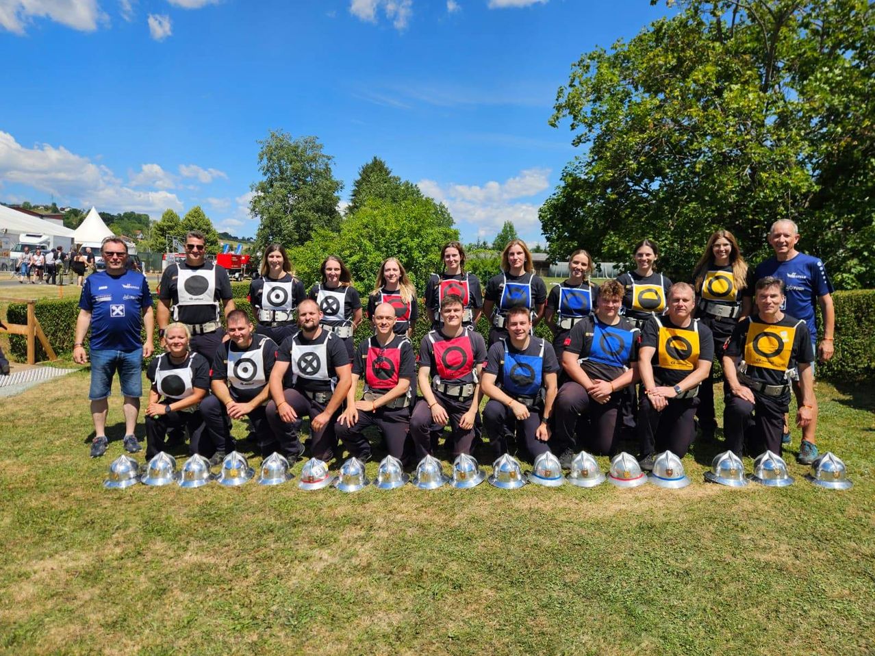 Eine Gruppe von Menschen in Sicherheitswesten mit Zieldesigns posiert für ein Foto auf einem Grasfeld. Vor ihnen stehen mehrere silberne Helme. Dahinter sind Bäume und ein blauer Himmel mit Wolken.