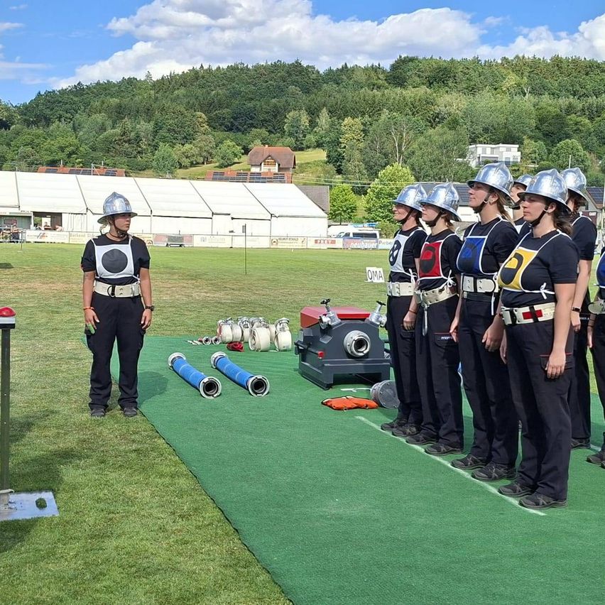 Eine Gruppe von Frauen in Helmen und Uniformen steht auf einem grünen Feld mit Ausrüstung in der Nähe. Eine Frau vorne trägt eine Schutzweste. Im Hintergrund sind ein großes weißes Zelt und ein Haus sichtbar.