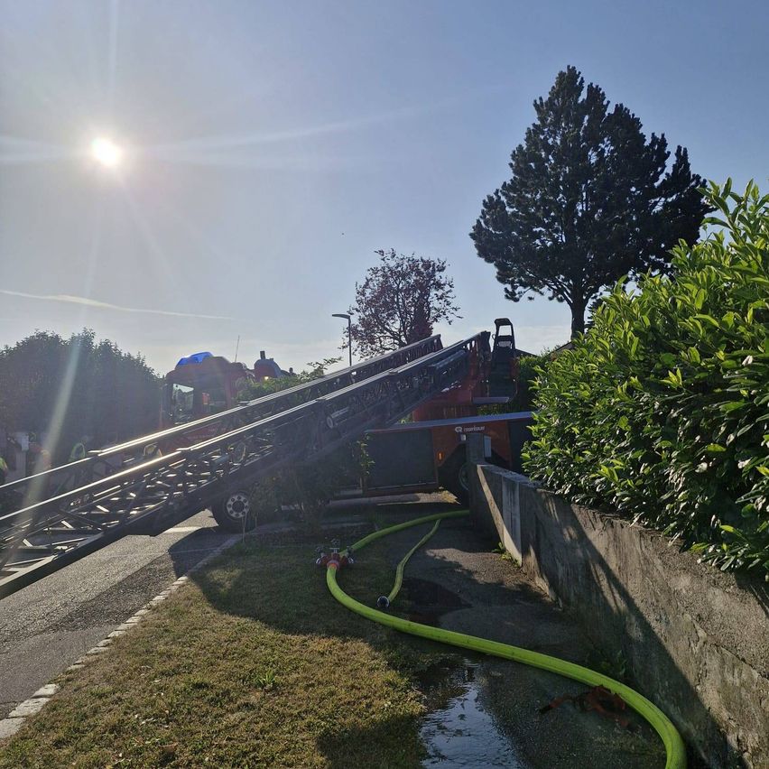 A fire truck with its ladder extended rests beside a wall, near a bush and trees, under a bright sky.
