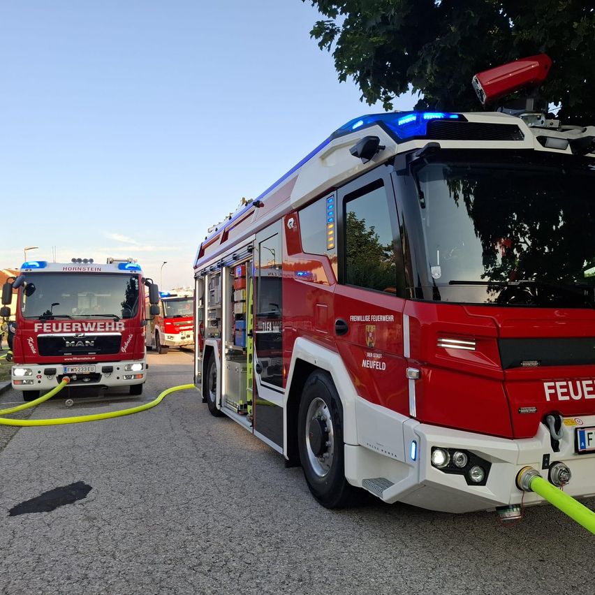 Zwei Feuerwehrfahrzeuge sind mit einem Schlauch verbunden auf der Straße geparkt. Bäume und Straßenlaternen sind im Hintergrund.