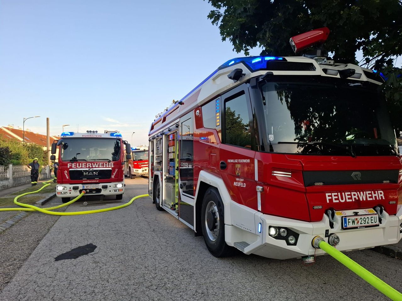 Two fire trucks are parked on the road with a hose connected. Trees and streetlights are in the background.
