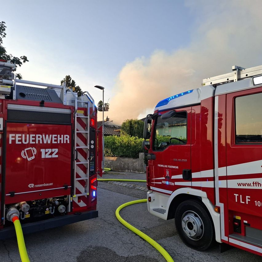 Zwei Feuerwehrfahrzeuge mit der Aufschrift 'Feuerwehr 122' und 'TLW 10' sind geparkt. Ein gelber Schlauch ist an einem der Fahrzeuge angeschlossen. Bäume und ein Gebäude befinden sich dahinter. Rauch steigt in den Himmel auf.