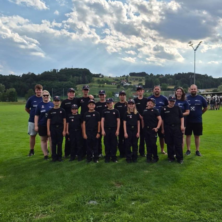 Eine Gruppe von Menschen in einheitlicher Kleidung, einschließlich Kindern, posiert für ein Foto auf einem Rasenfeld mit bewölktem Himmel und Hügeln im Hintergrund.