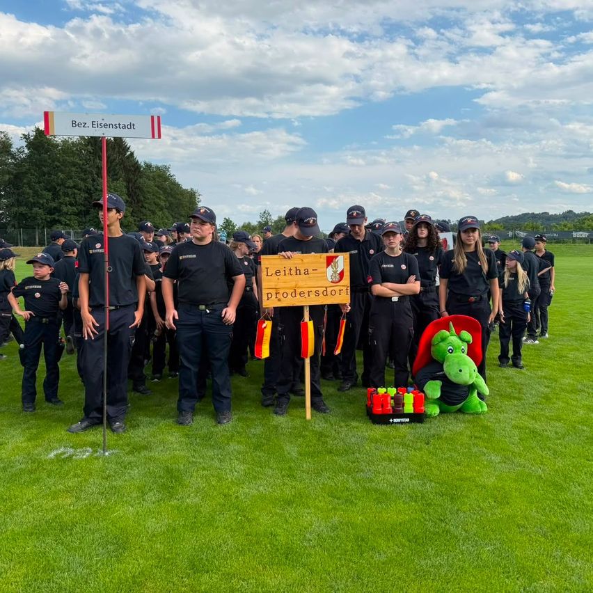 Eine Gruppe von Menschen in Uniform steht auf einem grünen Feld mit einem Schild, das 'Leithausproersdorf' und eine Maskottchen zeigt.