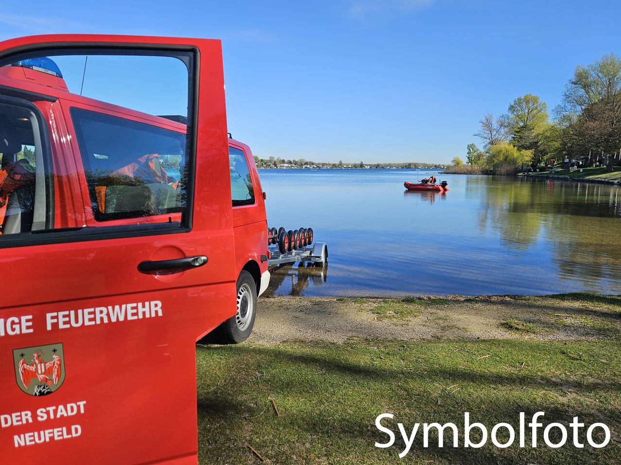 Ein roter Feuerwehrwagen steht in der Nähe eines Sees. Der Wagen hat die Worte 'Feuerwehr' und 'Stadt Neufeld' darauf geschrieben. Ein kleines Boot ist auf dem Wasser.