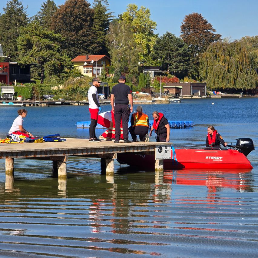 Eine Gruppe von Menschen steht auf einem Steg in der Nähe eines Sees. Ein rotes Boot ist im Wasser und eine Person sitzt darin.