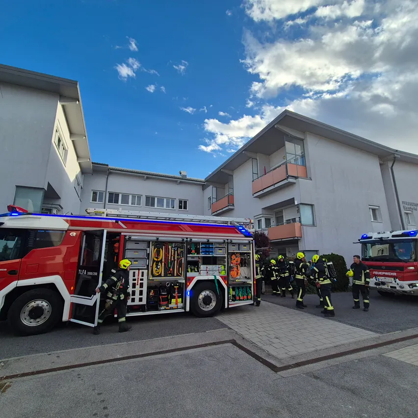 Eine Gruppe von Feuerwehrleuten steht vor einem Feuerwehrauto mit offener Tür. Das Feuerwehrauto ist vor einem Gebäude mit Balkonen geparkt.