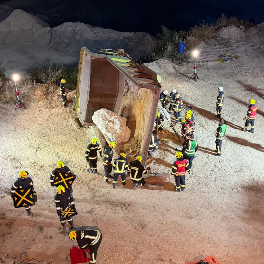 Eine Gruppe von Feuerwehrleuten in Schutzausrüstung versammelt sich um einen umgestürzten Lastwagen auf einer unbefestigten Straße bei Nacht.