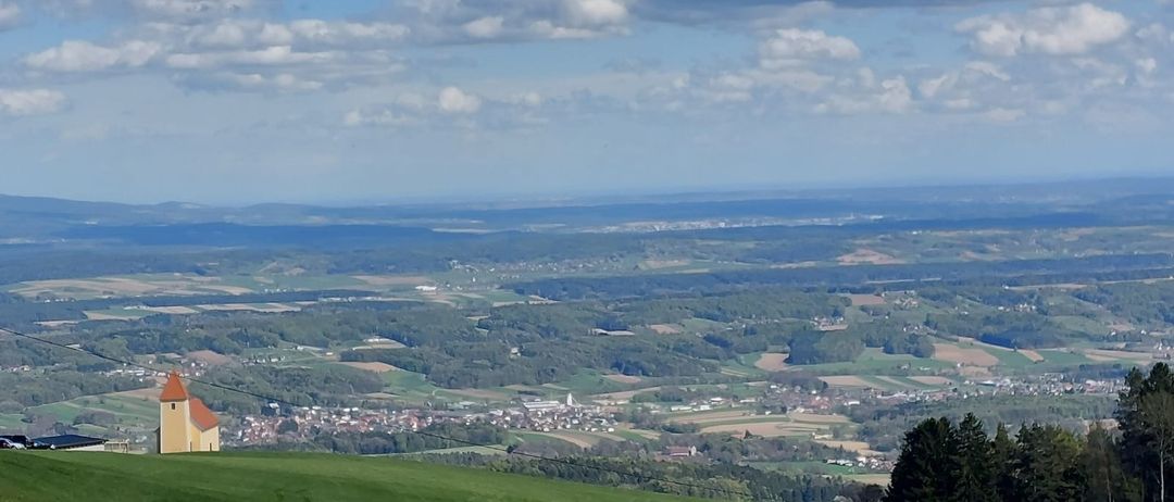 Luftaufnahme eines grünen Hügels mit einer kleinen Kapelle oben und einer Stadt unten unter einem bewölkten Himmel.