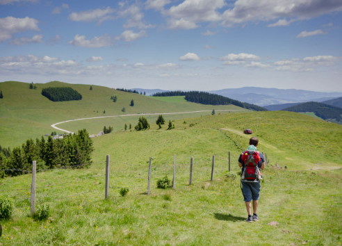 Eine Person mit einem Rucksack geht auf einem Grashügel, mit Blick auf eine kurvenreiche Straße mit entfernten Bergen und einem blauen Himmel mit Wolken.
