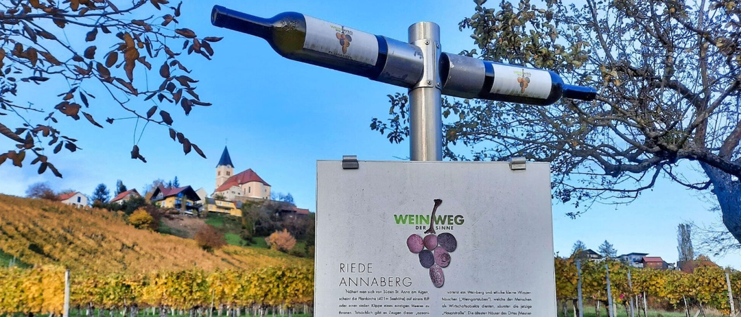 Ein Flaschenschild ist vor einem Weinbergshintergrund ausgestellt. Das Schild trägt die Aufschrift 'Reite Annaberg' und enthält Informationen über einen Weinberg und Weinverkostungen.