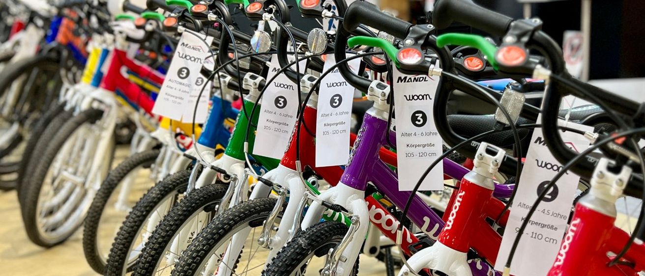 Ein Nahaufnahme von Kinderfahrrädern mit Preisschildern, die Größen und Altersempfehlungen zeigen. Die Fahrräder sind aufgereiht, in verschiedenen Farben und Marken, einschließlich Woom.