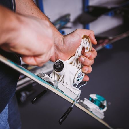 Eine Person benutzt ein Werkzeug, um die weißen Bindungen eines Snowboards zu justieren. Das Snowboard liegt auf einer dunklen Oberfläche.