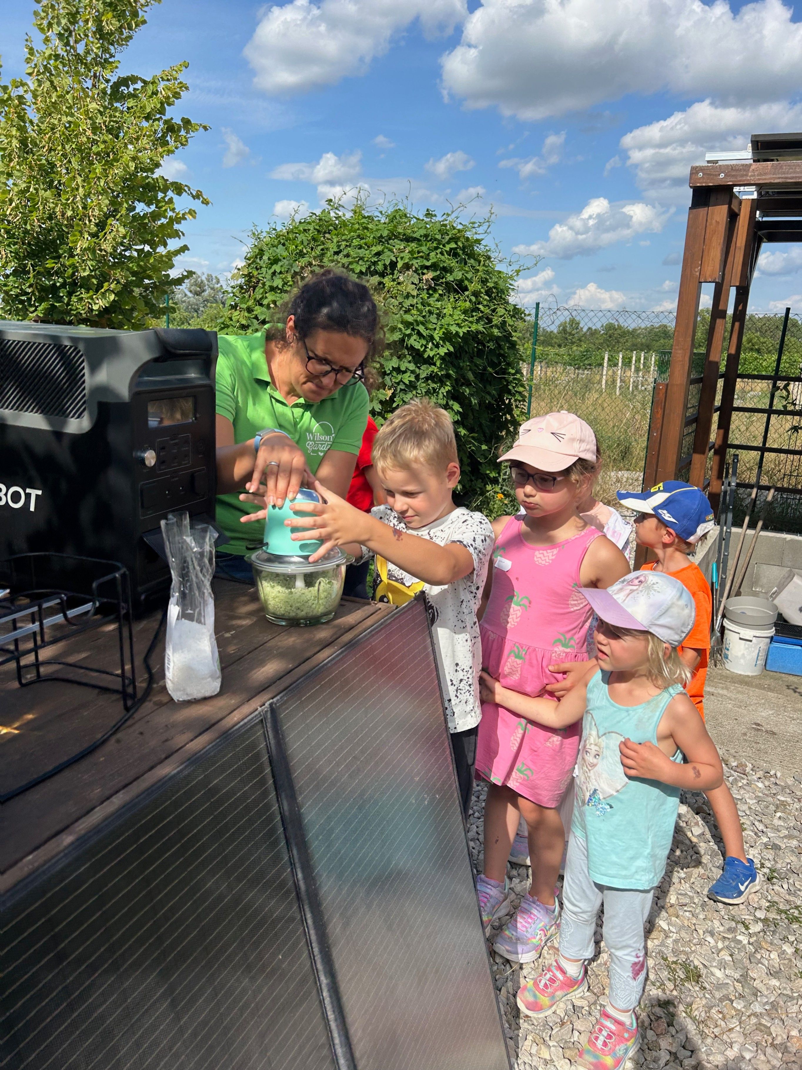 Eine Frau in einem grünen Shirt benutzt einen solarbetriebenen Mixer, um Essen zuzubereiten, während vier Kinder zusehen.