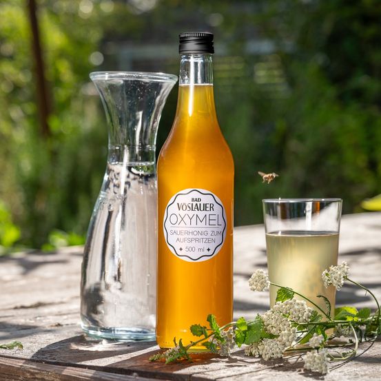 Eine Flasche 'Bad Voslauer Oxymel' wird auf einer Holzoberfläche mit einem Glaskrug und einem Glas mit einer Biene darauf ausgestellt. Die Flüssigkeit in der Flasche ist gelb und eine Pflanze mit weißen Blüten befindet sich rechts.