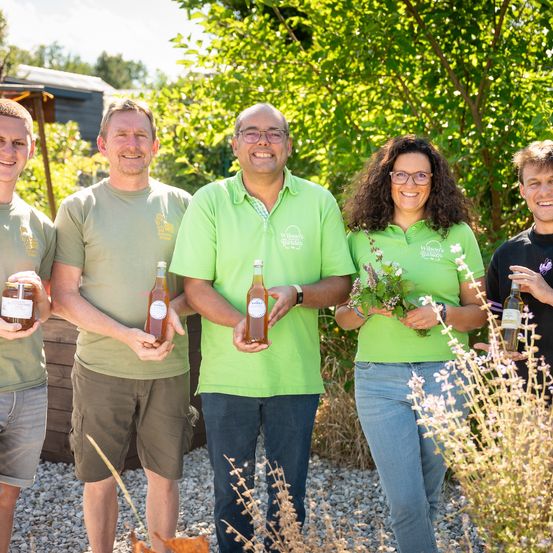 Fünf Menschen in grünen Shirts, einer hält eine Dose und ein anderer eine Flasche, stehen draußen mit Pflanzen und einem Gebäude dahinter.
