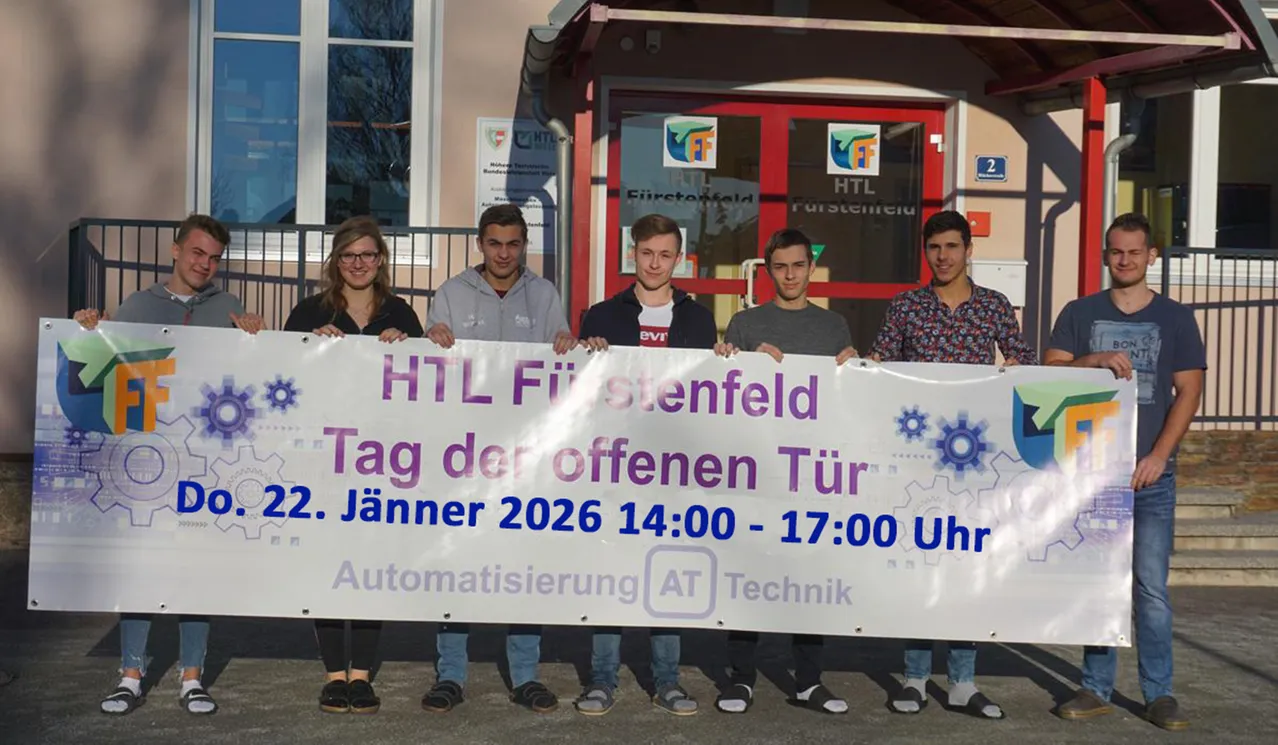 Fünf Personen stehen vor einem Gebäude und halten ein Banner. Das Banner trägt den Text 'HTL Furstenfeld Tag der offenen Tür 22. Jänner 2026 14:00-17:00 Uhr Automatisierung AT Technik'.