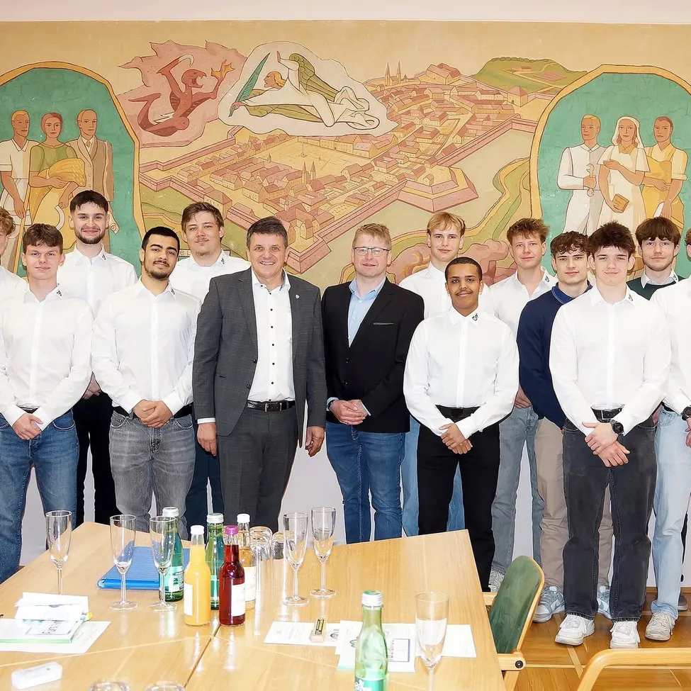 Eine Gruppe junger Männer und zwei Männer in Anzügen posieren für ein Foto vor einer Wand mit einem Wandbild. Sie stehen neben einem Tisch mit Flaschen, Gläsern und anderen Gegenständen.