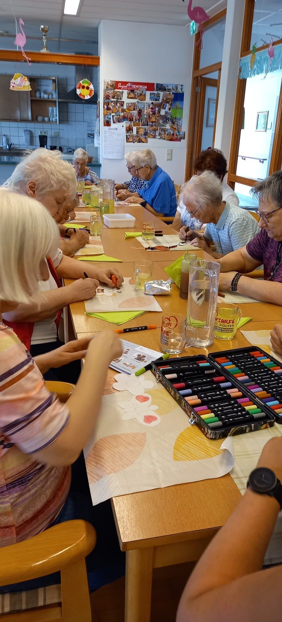 Eine Gruppe älterer Personen sitzt um einen Tisch herum und ist mit einer Malaktivität beschäftigt. Sie haben Malbücher, Marker und Gläser mit Wasser auf dem Tisch.