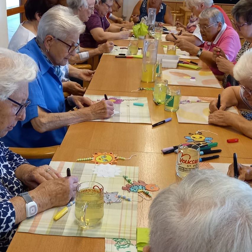 Eine Gruppe älterer Frauen sitzt um einen Tisch herum und ist in einer Malaktivität beschäftigt. Jede Person hat ein Malbuch und Marker vor sich. Der Tisch ist mit verschiedenen Gegenständen wie Gläsern mit Saft, Stiften und kleinen dekorativen Gegenständen gefüllt.