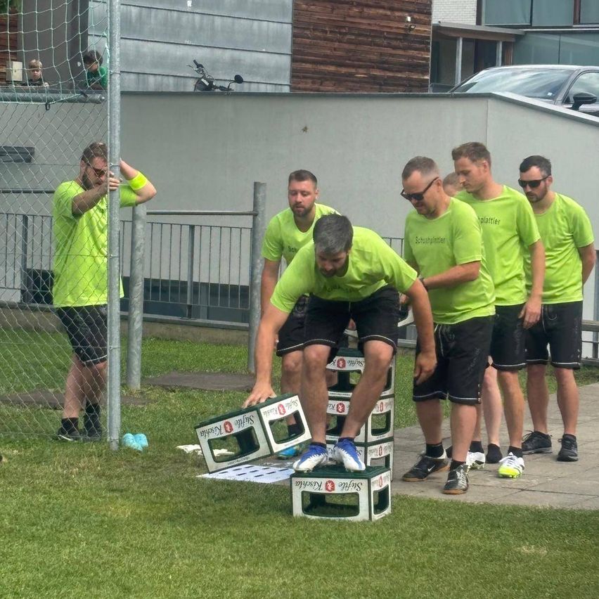Eine Gruppe von Männern in grünen Shirts und schwarzen Shorts steht auf einem Rasen. Ein Mann springt auf einen Stapel Kisten, während andere zuschauen. Dahinter befindet sich ein Zaun, ein Gebäude und ein geparktes Auto.