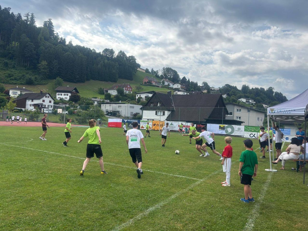 Eine Gruppe von Menschen spielt Fußball auf einem Feld, mit mehreren Häusern und Bäumen im Hintergrund. Der Himmel ist bewölkt.