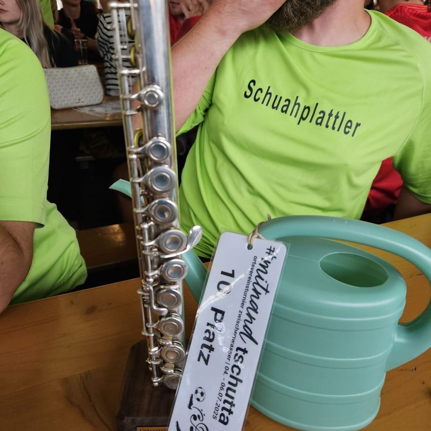 Eine Flöte wird auf einem Holztisch neben einem grünen Gießkanne ausgestellt. Ein Mensch in einem grünen Shirt sitzt hinter der Flöte. Das Shirt trägt Schuahplattler.