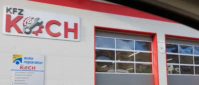 Ein weißes Gebäude mit rotem Rand hat zwei Garagentore. Das Schild zeigt KFZ Koch. Ein kleineres Schild hat die Wörter Auto Reparatur.