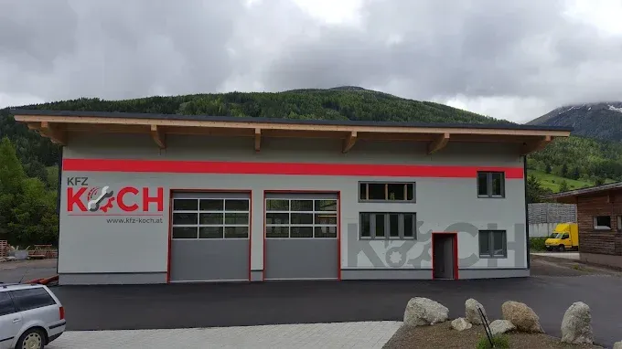 Ein graues Gebäude mit den Worten KFZ KOCH, zwei Garagen und einem kleinen Parkplatz. Im Hintergrund befinden sich Berge, ein gelber Van und ein kleines Gebäude.
