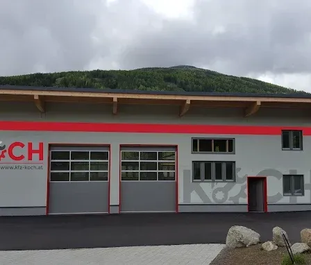 Ein graues Gebäude mit den Worten KFZ KOCH, zwei Garagen und einem kleinen Parkplatz. Im Hintergrund befinden sich Berge, ein gelber Van und ein kleines Gebäude.