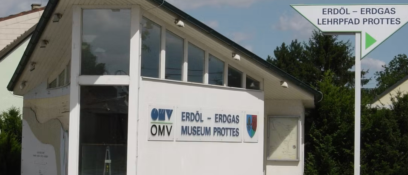 Ein Gebäude mit Glasfenstern und einer Bank davor. Es hat zwei Schilder, eines mit OMV und das andere mit Erdöl-Erdgas Museum Prottes.