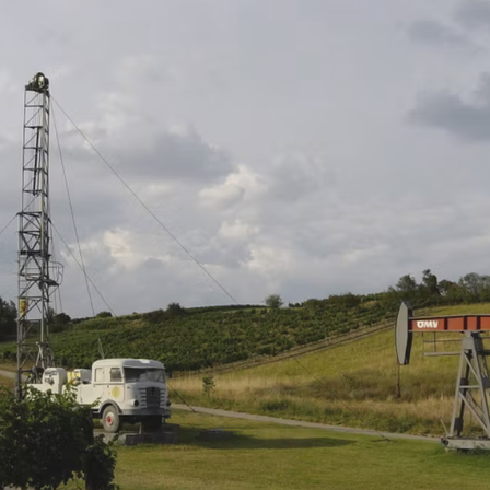 Ein alter Bohrturm steht auf einem grünen Feld unter einem bewölkten Himmel, mit einem kleinen Hügel, der von grünen Bäumen bedeckt ist.