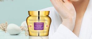 Eine Frau mit einem weißen Handtuch auf dem Kopf trägt Creme auf ihr Gesicht auf. Die Creme befindet sich in einem goldenen Glas mit Etikett. Sie schaut in die Kamera.