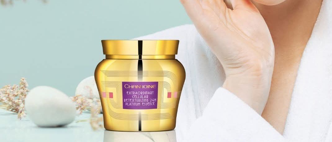 Eine Frau mit einem weißen Handtuch auf dem Kopf trägt Creme auf ihr Gesicht auf. Die Creme befindet sich in einem goldenen Glas mit Etikett. Sie schaut in die Kamera.