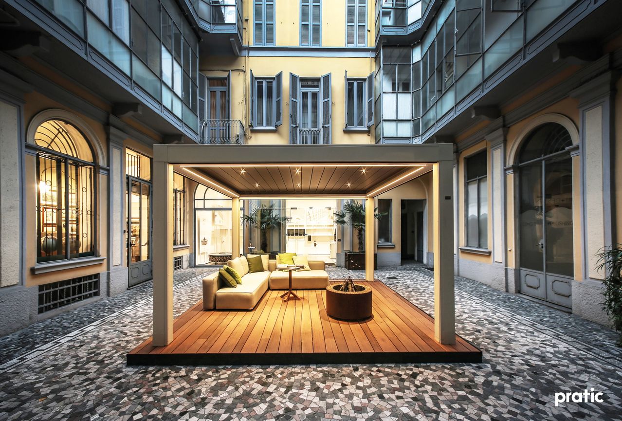 Ein Innenhof mit Holzboden, einem Sofa mit Kissen, einem kleinen Tisch und einem Feuerkorb. Der Bereich ist von Glaswänden umgeben und hat ein modernes architektonisches Design.