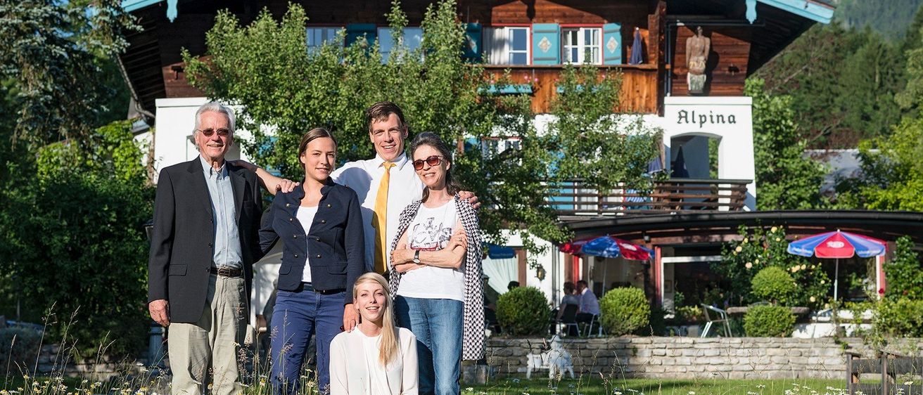 Fünf Personen posieren für ein Foto vor einem Gebäude mit der Aufschrift Alpina, umgeben von Bäumen und Blumen.