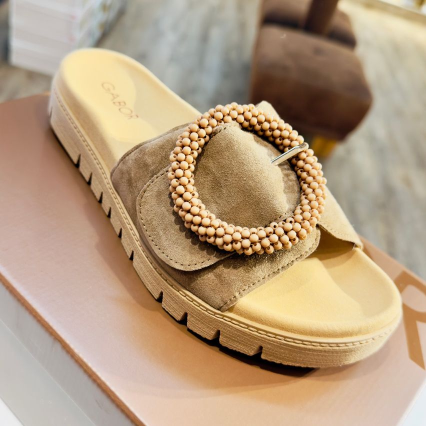 Ein Paar beigefarbener Sandalen mit einer perlenbesetzten Schnalle am Riemen wird auf einer braunen Schachtel ausgestellt. Der Markenname 'GABO' ist an der Sohle zu erkennen.
