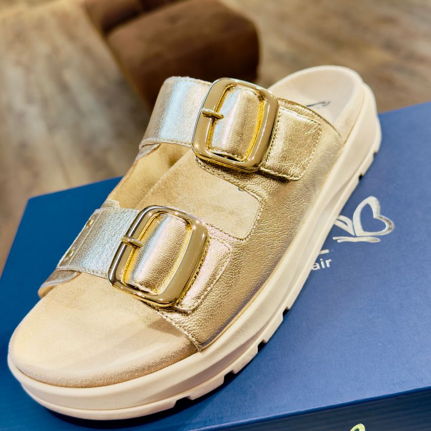 Ein Paar goldener Sandalen mit doppelten Riemen und Schnallen an weißer Sohle, auf einem blauen Karton präsentiert.
