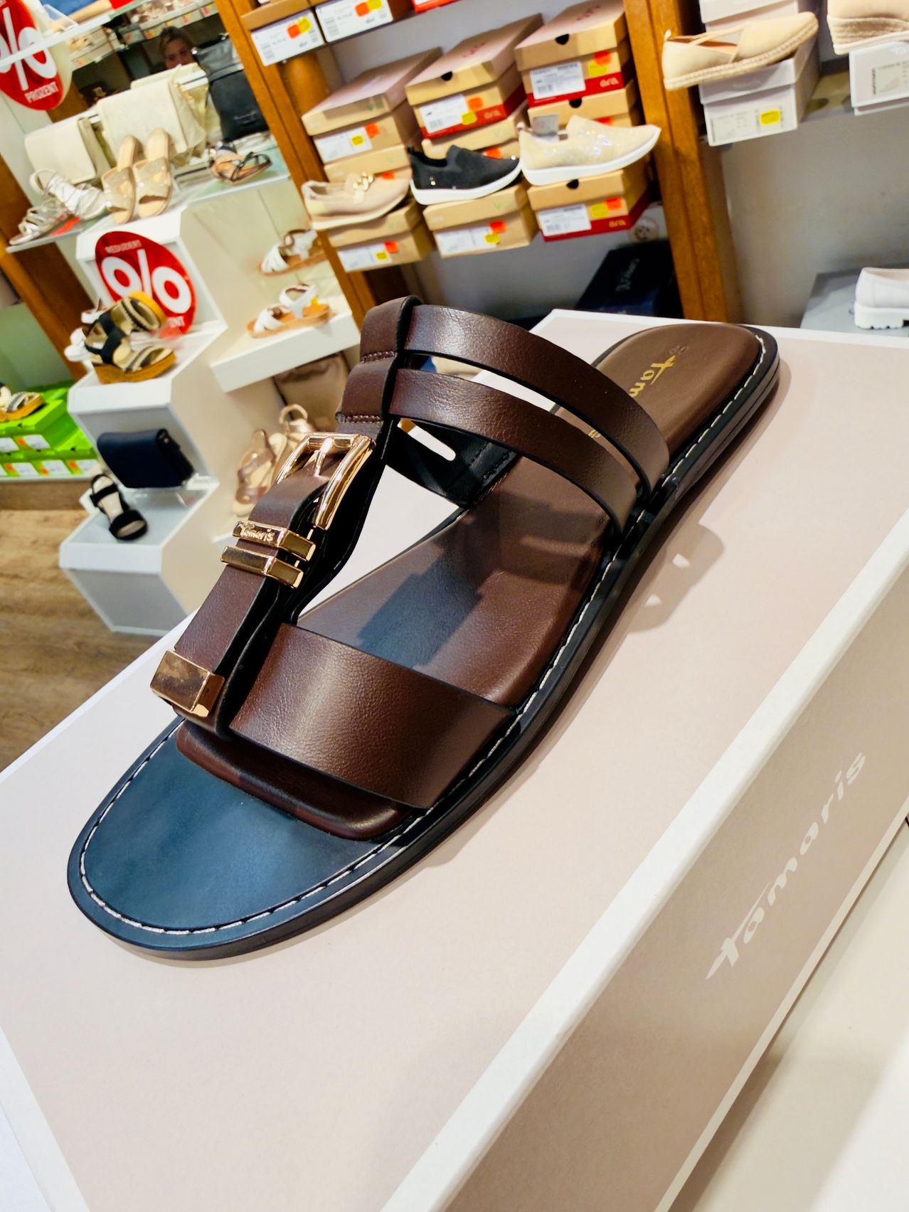 A brown leather sandal with gold buckles is displayed on a white box in a shoe store.