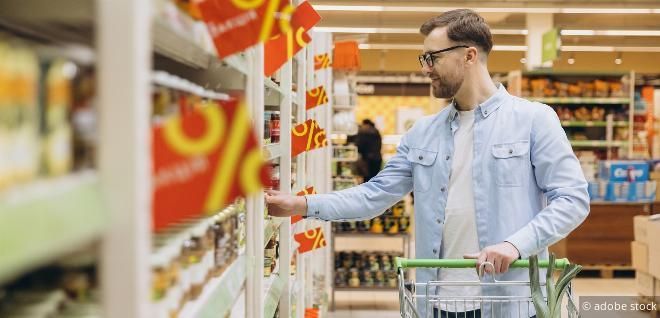 Ein Mann mit Brille und hellblauem Hemd durchstöbert Artikel in einem Supermarktregal und hält einen Einkaufswagen. Rabattschilder hängen an den Regalen.